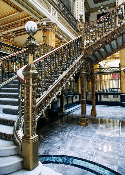 Escalier d'honneur en fer forgé doré et sol de marbre dans le hall d'un établissement bancaire ancien.
