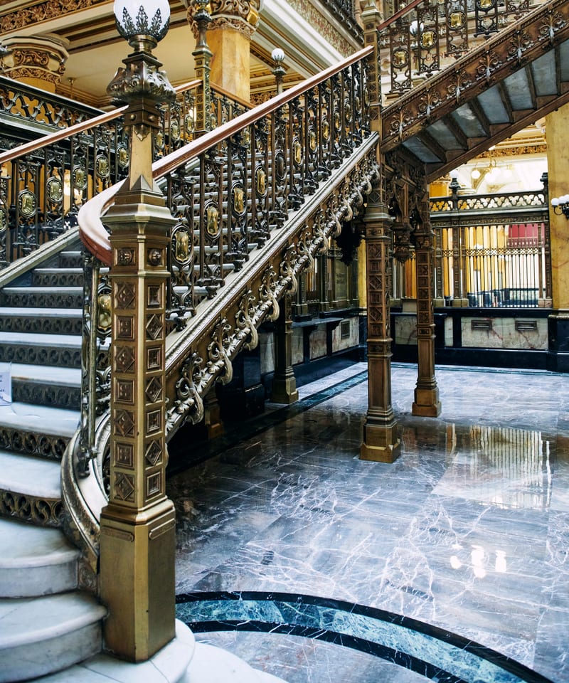Escalier d'honneur en fer forgé doré et sol de marbre dans le hall d'un établissement bancaire ancien.